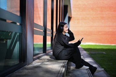 Genç kız açık havada kulaklıklarla oturmuş akıllı telefonuyla parlak güneş ışığı altında müzik dinliyor..