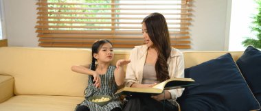 Mutlu Asyalı anne ve küçük kız kanepede masal vakit geçirmek, gülümseme, sevgi ve aile birlikteliği paylaşmak.