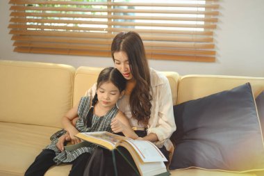 Mutlu Asyalı anne ve küçük kız kanepede masal vakit geçirmek, gülümseme, sevgi ve aile birlikteliği paylaşmak.