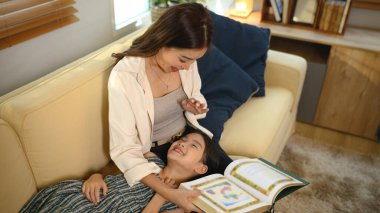 Bir anne, kızına kitap okuyor oturma odasında kucağında dinleniyor, huzurlu bir aile anının tadını çıkarıyor..