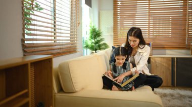 Asyalı bir anne ve kızı evde oturup kitap okuyup gülüyor ve kaynaşıyor..