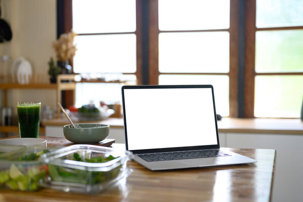 Ноутбук с чистым белым экраном на деревянном кухонном столе рядом с зеленым смузи и здоровой пищи.