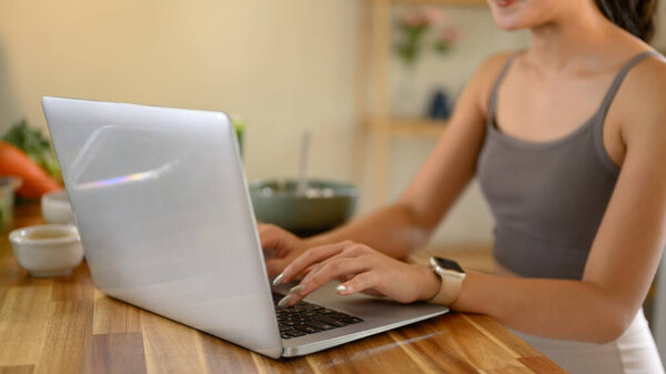 Крупный план женщины, печатающей на ноутбуке, сидящей за деревянным кухонным столом.