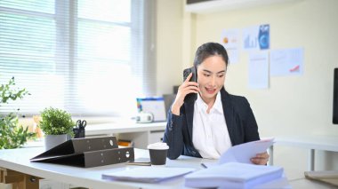 İş kadını masasındaki belgeleri incelerken telefonda konuşuyor, müşteri danışmanlığını gösteriyor..