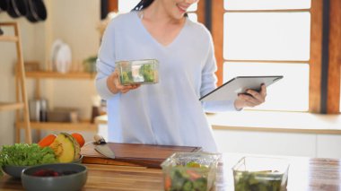 Mutfakta sağlıklı yemekler hazırlarken tablet kullanan gülümseyen kadın, yaşam tarzı dengesini ve ev yemeği pişirme ilhamını gösteriyor..