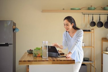 Mutfakta sağlıklı yemekler hazırlarken tablet kullanan gülümseyen kadın, yaşam tarzı dengesini ve ev yemeği pişirme ilhamını gösteriyor..