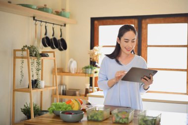Mutfakta sağlıklı yemekler hazırlarken tablet kullanan gülümseyen kadın, yaşam tarzı dengesini ve ev yemeği pişirme ilhamını gösteriyor..