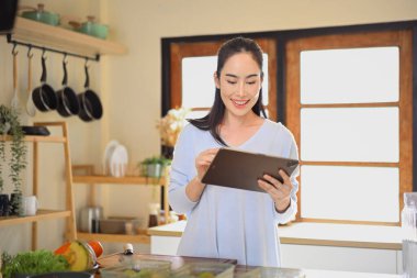 Mutfakta sağlıklı yemekler hazırlarken tablet kullanan gülümseyen kadın, yaşam tarzı dengesini ve ev yemeği pişirme ilhamını gösteriyor..