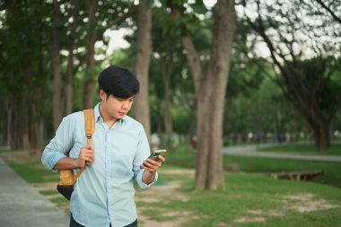 Telefonunu kontrol ederken parkta yürüyen bir adam, rahat bir şekilde dışarıda sakin bir anın tadını çıkarıyor..