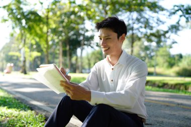 Bir adam yere oturmuş defterine notlar yazıyor, parkta huzurlu bir anın tadını çıkarıyor..