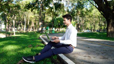 Parktaki bir dizüstü bilgisayarla çimlerin üzerinde oturan bir adam, sakin ve doğal bir açık havada çalışıyor..