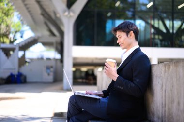 Ofisteki adam elinde kahve fincanıyla dışarıda dizüstü bilgisayarda çalışıyor..