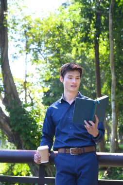 Ofisten bir adam elinde kahve fincanı tutarken gölün kenarında durup kitap okuyor..