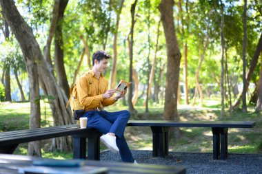 Yeşil bir parkta bankta oturan bir adam kahve fincanıyla kitap okuyor..