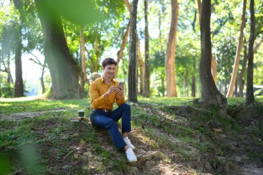 Ofisten bir adam yeşil bir parktaki çimlerin üzerinde rahat bir şekilde telefonunu kullanıyor..