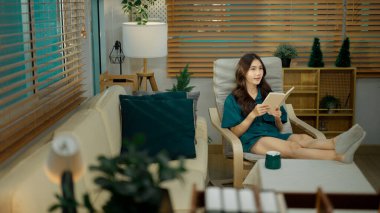 Oturma odasındaki koltukta dinlenirken kitap okuyan kadın..