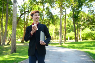 Parlak bir günde elinde laptop ve kahve fincanı olan bir adam yeşil bir parkta yürüyor..