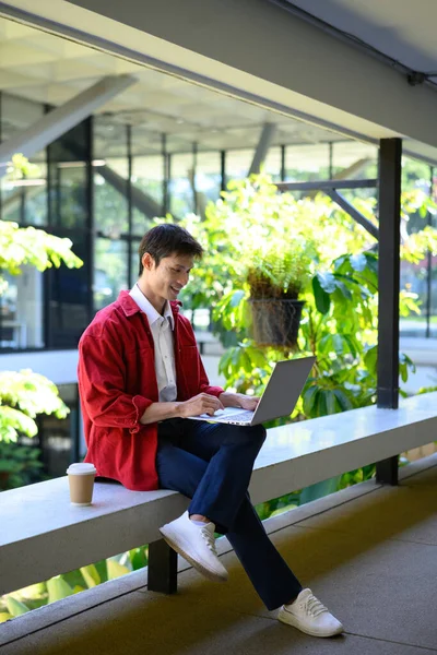 Profesyonel bir adam, modern bir binanın dışında dizüstü bilgisayarla çalışmaya odaklanmış..