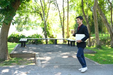 Parlak bir günde elinde laptop ve kahve fincanı olan bir adam yeşil bir parkta yürüyor..