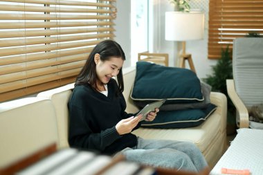 Genç bir kadın evinde tablet kullanarak kanepede dinleniyor, rahat ve sakin bir tatil havasının tadını çıkarıyor..