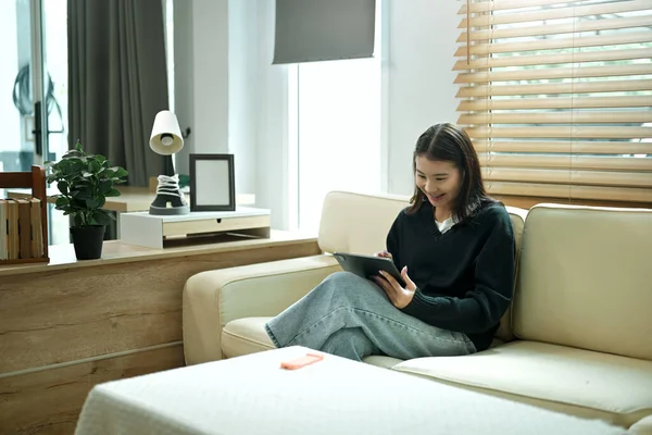 Genç bir kadın evinde tablet kullanarak kanepede dinleniyor, rahat ve sakin bir tatil havasının tadını çıkarıyor..