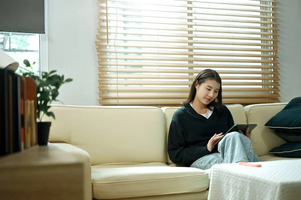 Genç bir kadın evinde tablet kullanarak kanepede dinleniyor, rahat ve sakin bir tatil havasının tadını çıkarıyor..