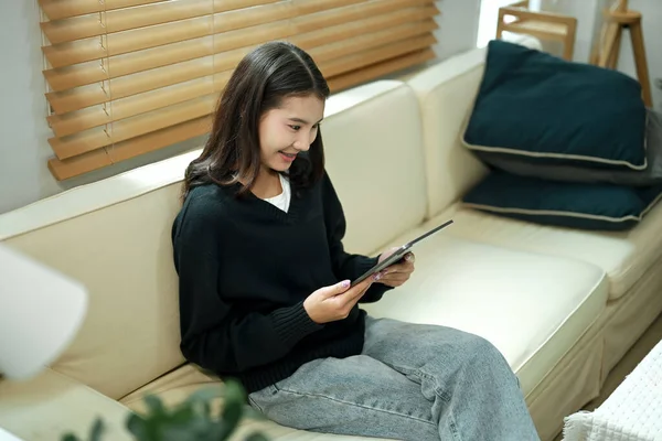 Genç bir kadın evinde tablet kullanarak kanepede dinleniyor, rahat ve sakin bir tatil havasının tadını çıkarıyor..