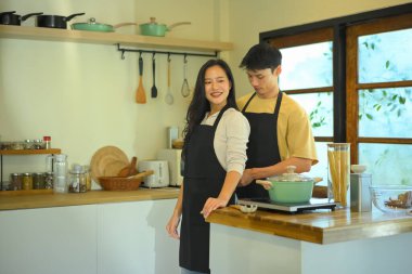 Genç Asyalı çift evde birlikte yemek pişiriyor, gülümsüyor erkek de kadın önlüğünün rahat, güneşli bir mutfakta ayarlanmasına yardım ediyor...