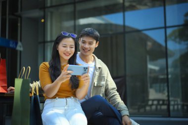 Alışveriş yaptıktan sonra dışarıda dinlenen Asyalı çift renkli alışveriş torbalarının yanında akıllı telefonları kontrol ederken gülümsüyor..