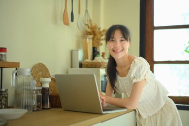 Genç bir kadın mutfakta dizüstü bilgisayarla çalışırken ve mutfakta kameraya bakarken..