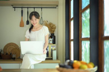 Genç bir kadın mutfak tezgahında dizüstü bilgisayar kullanıyor..