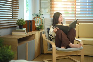 Happy relaxed young woman resting on armchair reading book and holding coffee cup.