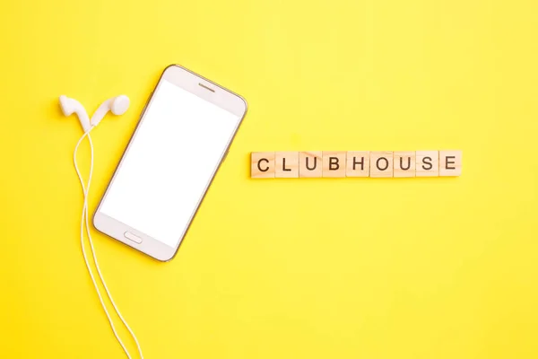 Smartphone et écouteurs et blocs de bois avec des lettres forment le mot clubhouse médias sociaux sur un fond jaune. Concept de chat audio. Photos De Stock Libres De Droits