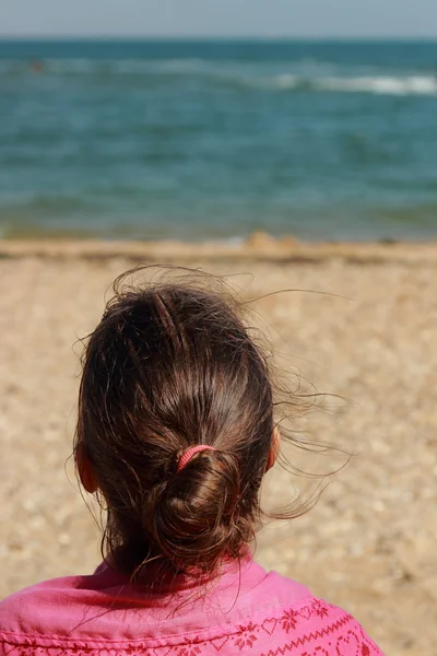 Küçük kız kumsalda oturuyor. Sırtı kameraya dönük. Yazın güneşli bir günde.