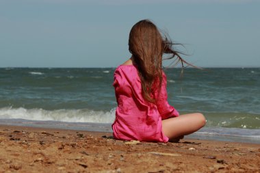 Küçük kız kumsalda oturuyor. Sırtı kameraya dönük. Yazın güneşli bir günde.