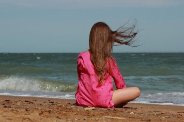 Küçük kız kumsalda oturuyor. Sırtı kameraya dönük. Yazın güneşli bir günde.