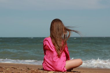 Küçük kız kumsalda oturuyor. Sırtı kameraya dönük. Yazın güneşli bir günde.