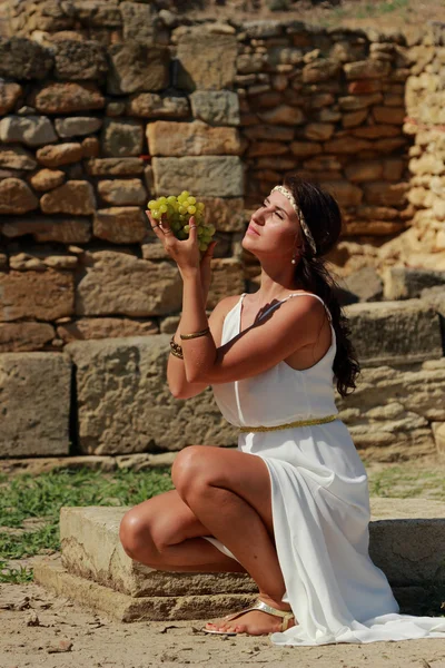 在希腊古代城市科尔奇的挖掘背景下 有着漂亮的希腊风格头发的迷人的小女孩吃着白葡萄 — 图库照片