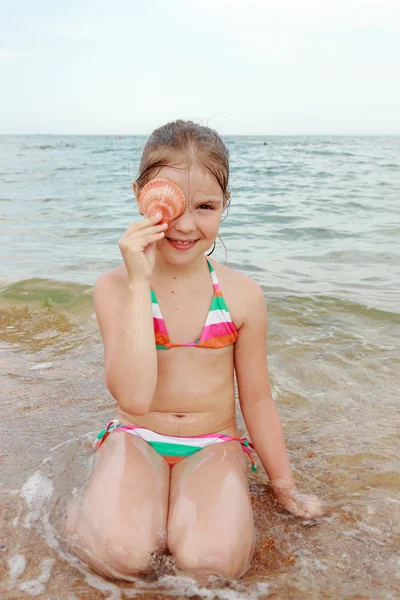 海と夏の日の貝殻を持って座っている明るい水着で幸せな陽気な少女 — ストック写真