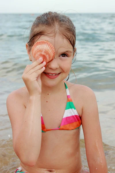 海と夏の日の貝殻を持って座っている明るい水着で幸せな陽気な少女 — ストック写真