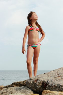 Tema yaz tatil ve seyahat sahilde deniz arka plan ve mavi gökyüzü üzerinde büyük bir kayanın üzerinde bekleyen bir mayo içinde mutlu küçük kız