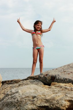 Tema yaz tatil ve seyahat sahilde deniz arka plan ve mavi gökyüzü üzerinde büyük bir kayanın üzerinde bekleyen bir mayo içinde mutlu küçük kız