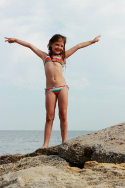 Tema yaz tatil ve seyahat sahilde deniz arka plan ve mavi gökyüzü üzerinde büyük bir kayanın üzerinde bekleyen bir mayo içinde mutlu küçük kız