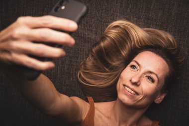 Happy mid-aged woman taking selfie with her smart phone at home clipart
