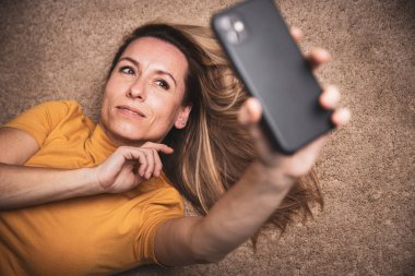 Mutlu orta yaşlı kadın evde akıllı telefonuyla selfie çekiyor.