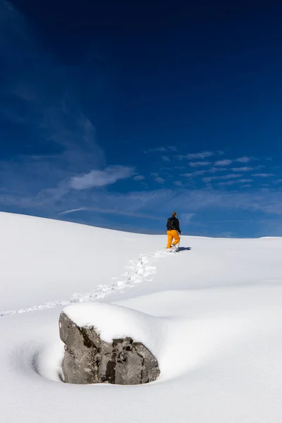冬のスポーツ 多くの雪で覆われた高い山の中で上り坂のスノーシューで歩く若い男 背景の山に選択的な焦点 — ストック写真