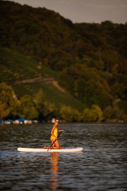 Suyun altından çekilen sıcak öğleden sonra ışığında güzel, genç bir kadın güzel bir gölde kürek çekiyor.