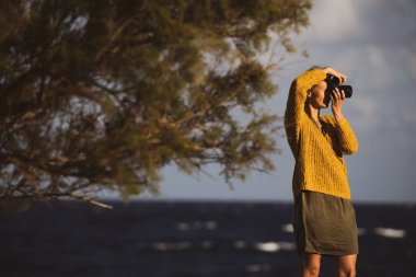 Aynasız kamerası olan güzel, genç bir kadın, deniz kenarında fotoğraf çekiyor.