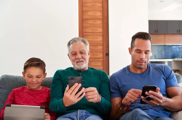 携帯電話やデジタルタブレットを使用して自宅でソファに座っている世代の男性ヒスパニック系家族 — ストック写真