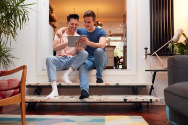 Loving Same Sex Male Couple Using Digital Tablet As They Relax At Home Together clipart